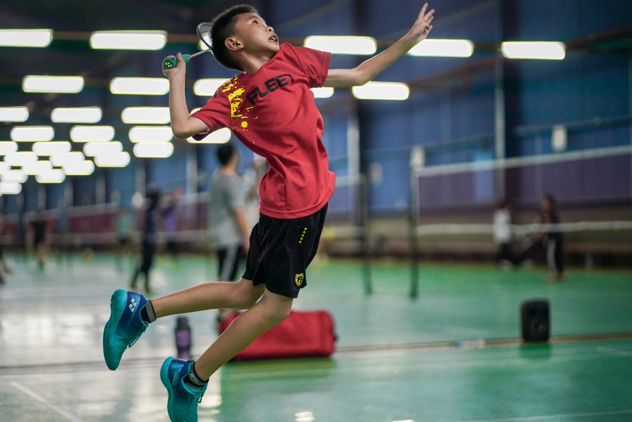 our-services-02 A young boy enthusiastically jumps to hit a badminton shuttlecock inside a sports hall.